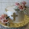 The RC-001H is a round white cake with gold drip accents, topped with pink roses and baby's breath, featuring a gold decorative cross at the center, and presented on a gold floral-patterned cake board.