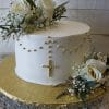 RC-002 is a white cake adorned with white roses, baby’s breath, greenery, and a gold bead rosary cross, displayed on a gold cake board against a white wooden background.