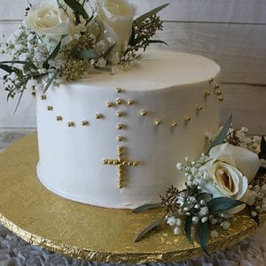 RC-002 is a white cake adorned with white roses, baby’s breath, greenery, and a gold bead rosary cross, displayed on a gold cake board against a white wooden background.