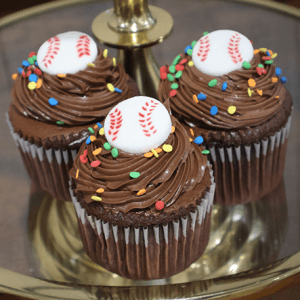 Three CC-096B chocolate cupcakes with white filling, chocolate frosting, colorful sprinkles, and white baseball-shaped toppers are displayed on a gold tray.