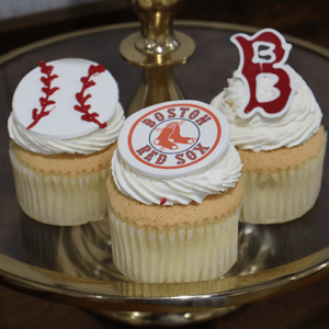 A CC-118A Gold cupcake with chocolate filling is shown on a gold cake stand, topped with edible baseball decorations, including the Boston Red Sox logo and a red letter B.