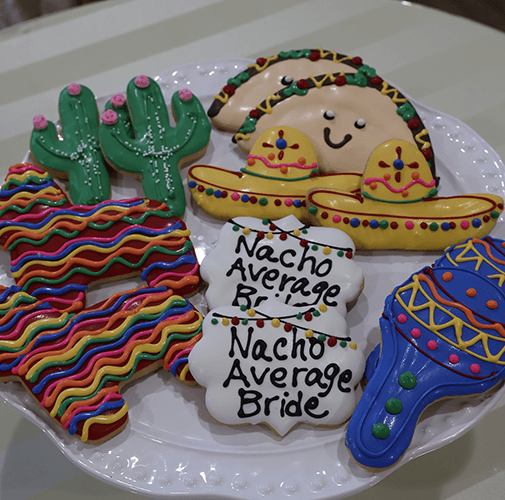 A plate of colorful CE-003i bridal sugar cookies shaped like cacti, tacos, sombreros, piñatas, maracas, and plaques reading “Nacho Average Bride.”.