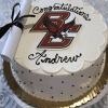 A white cake (GC-000B Boston College) features black polka dots, a Boston College logo, “Congratulations Andrew,” and a rolled-up paper tied with a black ribbon resting on top.