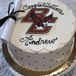 A white cake (GC-000B Boston College) features black polka dots, a Boston College logo, “Congratulations Andrew,” and a rolled-up paper tied with a black ribbon resting on top.