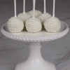 Six DB-000B Gold Cake Pops, each on a stick with gold coating and sprinkles, are arranged on a white beaded-edge cake stand, set on a marble surface against a dark brick background.