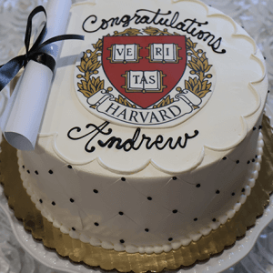 The GC-000E Harvard is a white graduation cake featuring a Harvard emblem, "Congratulations Andrew," black dots, and a small diploma tied with a black ribbon on top.