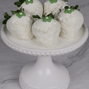 Five DB-000F Bride Strawberries, each dipped in white chocolate and decorated with icing and green floral details, are displayed on a white beaded-edge cake stand against a marble backdrop.