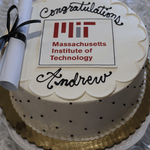 Round cake (Product: GC-000G MIT) with white icing, black dots, gold base. "Congratulations Andrew" on top with an MIT logo and diploma scroll tied with black ribbon, celebrating admission to Massachusetts Institute of Technology.