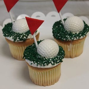 Three CC-116 Gold cupcakes with chocolate mousse filling are topped with white golf ball shapes, green grass-like sprinkles, and red flag decorations, arranged on a white scalloped-edge cake stand.