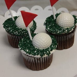 Three CC-115 chocolate cupcakes with white filling, decorated with green sprinkles, each topped with a white golf ball-shaped candy and a red triangular flag on a stick, displayed on a white stand.