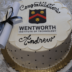 The GC-000J Wentworth is a white cake with black polka dots, topped with a rolled diploma and the Wentworth Institute of Technology logo. “Congratulations Andrew” is written in black icing.