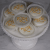 A DB-000H White Dipped Oreo sits in a white cake stand, with eight cookies in paper cups topped with gold sprinkles and yellow iced script, set against a soft gray marble background.