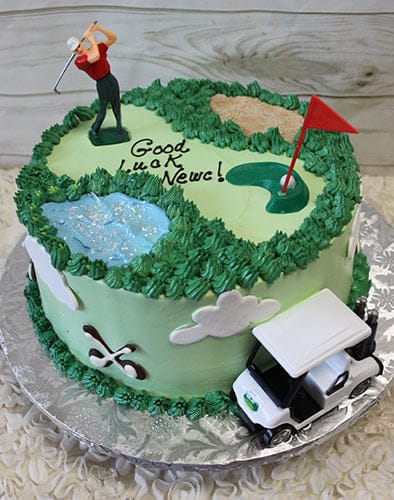 ST-007C Golf Scene: Round golf-themed cake with green frosting, golfer figurine, red flag, sand trap, water feature, "Good Luck Neve!" in icing, and a small toy golf cart at the base.