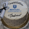 The GC-000K Bentley is a round white cake with "Congratulations Andrew" in black icing, the Bentley University logo in the center, a rolled-up diploma with a black ribbon on top, a gold base, and small dot decorations.