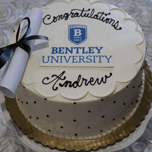 The GC-000K Bentley is a round white cake with "Congratulations Andrew" in black icing, the Bentley University logo in the center, a rolled-up diploma with a black ribbon on top, a gold base, and small dot decorations.