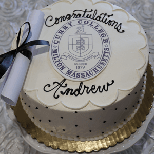 The GC-000M Curry College is a round white cake with black polka dots, featuring the Curry College seal, "Congratulations Andrew," and a rolled diploma tied with a black ribbon on top, served on a gold-edged platter.