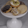 A white cake stand displays DB-000M Tea Cookies—an assortment topped with chocolate drizzle, powdered sugar, sliced almonds, and crushed pistachios—on a marble surface.