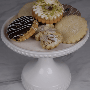 A white cake stand displays DB-000M Tea Cookies—an assortment topped with chocolate drizzle, powdered sugar, sliced almonds, and crushed pistachios—on a marble surface.