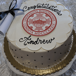 The GC-000D Boston University cake is round and white with black dots, topped with a BU seal, a scroll tied with a black ribbon, and "Congratulations Andrew" in black icing. It is presented on a gold tray.