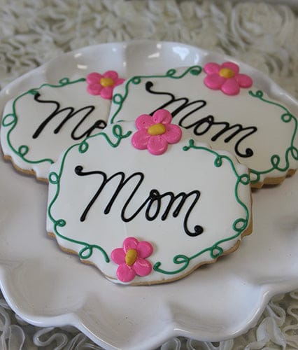 Three CE-050b Mother's Day cookies are on a white plate, each with "Mom" in black icing, green borders, and a pink flower with yellow center. The background is light and textured.