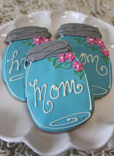 Three blue mason jar-shaped cookies from CE-050a Mother's Day, with "mom" in white icing and pink flower and green leaf decorations, are arranged on a white plate.