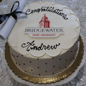 A white frosted GC-000R Bridgewater State University cake with “Congratulations Andrew,” the university logo in the center, and a rolled diploma with a black ribbon placed beside it.