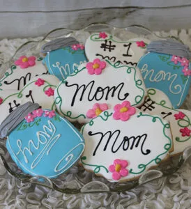 A plate of CE-050d Mother's Day cookies shaped like jars and plaques, decorated with “Mom,” “#1 Mom,” pink flowers, and blue or white icing in cursive.