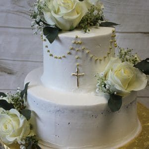 The RC-001A is a two-tier white cake with white roses, greenery, baby's breath, gold bead garlands, and a gold cross on the front. It's presented on a gold cake board against a wood-patterned background.