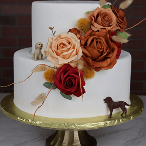 WD-011 is a two-tiered white cake adorned with orange, red, and peach flowers, dried grasses, and two small dog figurines, displayed on a gold stand against a brick backdrop.