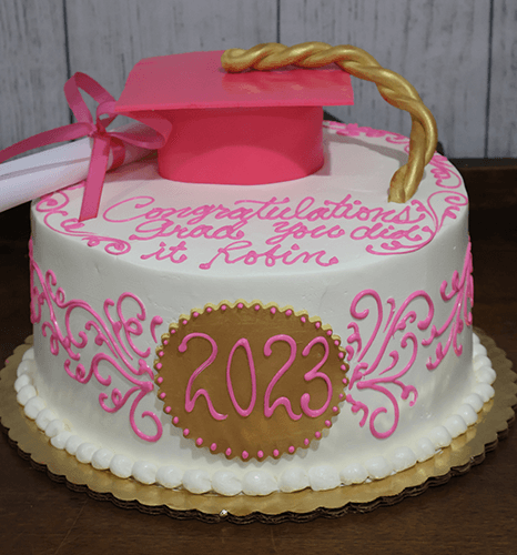 A round graduation cake (GC-019) with pink and gold icing, 2023 on the side, a pink grad cap topper, and “Congratulations Grad, You did it Robin” written on it.