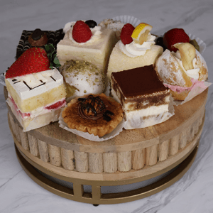 DB-000N Assorted Pastries: A selection of mini cakes and pastries, such as tiramisu, fruit-topped cakes, and nut tarts, beautifully arranged on a round wooden platter against a marble backdrop.