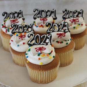 GC-070 Gold cupcakes with chocolate filling, decorated with white frosting, colorful sprinkles, and red bows, each topped with a black “2021” decoration, are arranged on a white cake stand.