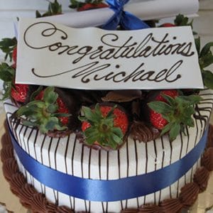 GC-002 is a decorated cake featuring chocolate-dipped strawberries, a blue ribbon, a congratulatory card for Michael in cursive, and a rolled-up paper tied with a blue bow on top.