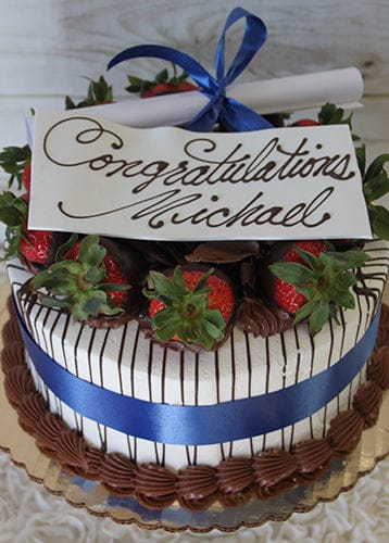GC-002 is a decorated cake featuring chocolate-dipped strawberries, a blue ribbon, a congratulatory card for Michael in cursive, and a rolled-up paper tied with a blue bow on top.