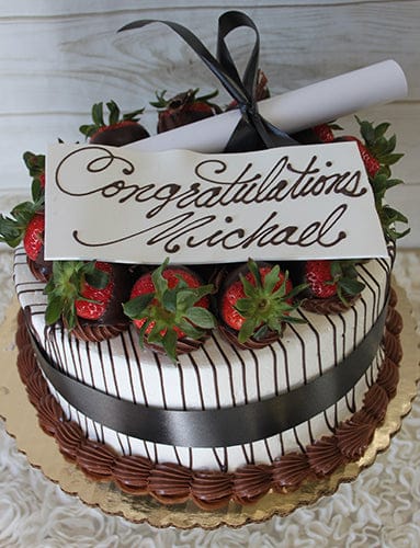 GC-003 is a round cake adorned with chocolate-dipped strawberries, chocolate drizzle, a black ribbon, and a scroll. A white plaque on top displays “Congratulations Michael” in elegant script.