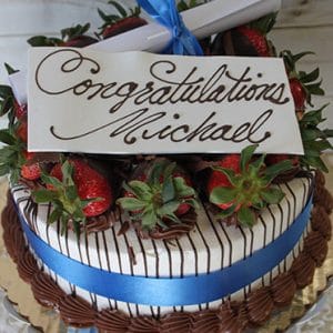 GC-005 is a decorated cake featuring chocolate-covered strawberries, a blue ribbon, and a white chocolate plaque reading "Congratulations Michael" in cursive, topped with a rolled-up paper tied with a blue ribbon.