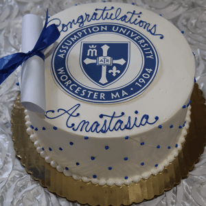 A round white cake on a gold board with the Assumption University Worcester MA logo, blue "Congratulations Anastasia" writing, and a diploma design. Product: GC-000T Assumption University.