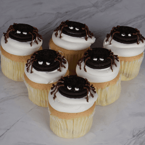 Seven CC-059 Spider cupcakes, gold with chocolate mousse filling, are topped with Oreo cookies decorated as spiders with chocolate legs and white eyes, arranged on a marble surface.