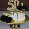 The Golden 50th cake features white icing with a gold drip, a large gold 50 topper, black fondant roses, gold leaves, and "Happy Birthday" on top, presented on a gold stand against a brick wall background.