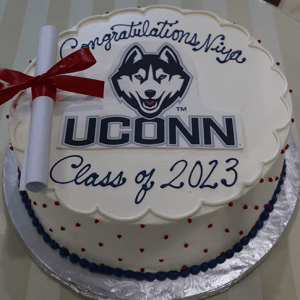 The GC-000U UConn cake features a round design with a UConn Husky logo, a red-ribboned diploma, and "Congratulations Niya Class of 2023" in blue icing. It is presented on a striped tablecloth.