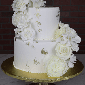 The TC-000B features a two-tiered white cake with gold leaf accents and large white flowers, presented on a gold cake stand in front of a brick wall.