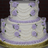 Product WD-002: Two-tier white cake with light purple frosting roses, white piped swags, green leaves, and purple borders on a gold board, set against a dark brick background.