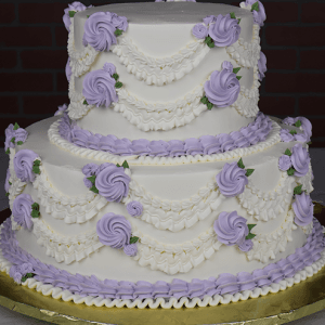Product WD-002: Two-tier white cake with light purple frosting roses, white piped swags, green leaves, and purple borders on a gold board, set against a dark brick background.