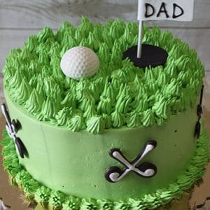 FD-011 is a round cake with green grass-like frosting, topped with a golf ball and a "#1 Dad" flag. Edible golf club decorations line the sides, and it is presented on a gold cake board.