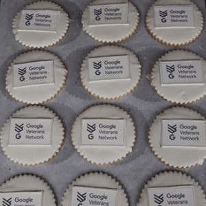 A tray of CO-120 Logo Cookies—round treats topped with white icing and the Google Veterans Network logo—are neatly arranged in rows on a baking sheet.