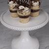 Three CO-101 Chocolate Peanut Butter Shooters, layered with cream and chocolate, topped with whipped cream and chocolate decorations, are displayed on a white cake stand against a marble background.