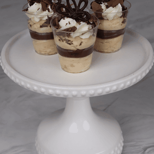 Three CO-101 Chocolate Peanut Butter Shooters, layered with cream and chocolate, topped with whipped cream and chocolate decorations, are displayed on a white cake stand against a marble background.