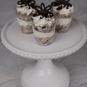 Three CO-105 Oreo Shooters, featuring layers of cream and cookie crumbles, topped with whipped cream, chocolate accents, and cookie crumbs, are displayed on a beaded-edge white cake stand.