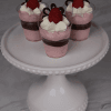 Three CO-102 Chocolate Raspberry Shooters—small dessert cups with pink mousse, whipped cream, raspberry, and chocolate decorations—are displayed on a white, beaded-edge cake stand against a marble background.