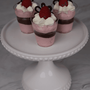 Three CO-102 Chocolate Raspberry Shooters—small dessert cups with pink mousse, whipped cream, raspberry, and chocolate decorations—are displayed on a white, beaded-edge cake stand against a marble background.
